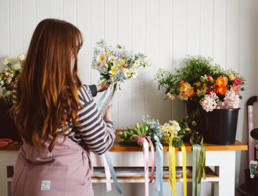 Maddie Jayde in her studio holding a pastel bouquet with trailing ribbons, wearing Collective Gen Charlie Overalls in Rose