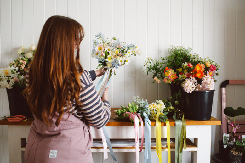 Maddie Jayde in her studio holding a pastel bouquet with trailing ribbons, wearing Collective Gen Charlie Overalls in Rose