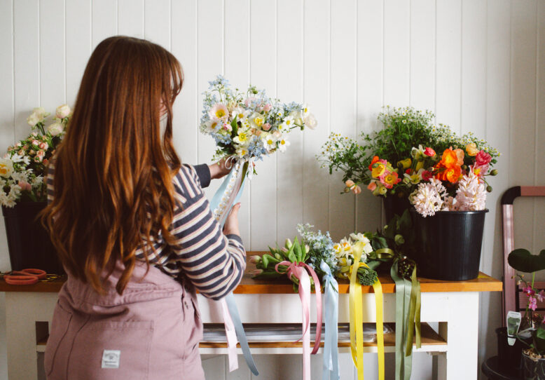 Maddie Jayde in her studio holding a pastel bouquet with trailing ribbons, wearing Collective Gen Charlie Overalls in Rose