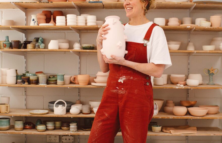 Brittney Kleyn smiling and holding a large pink vase in front of shelves of ceramics